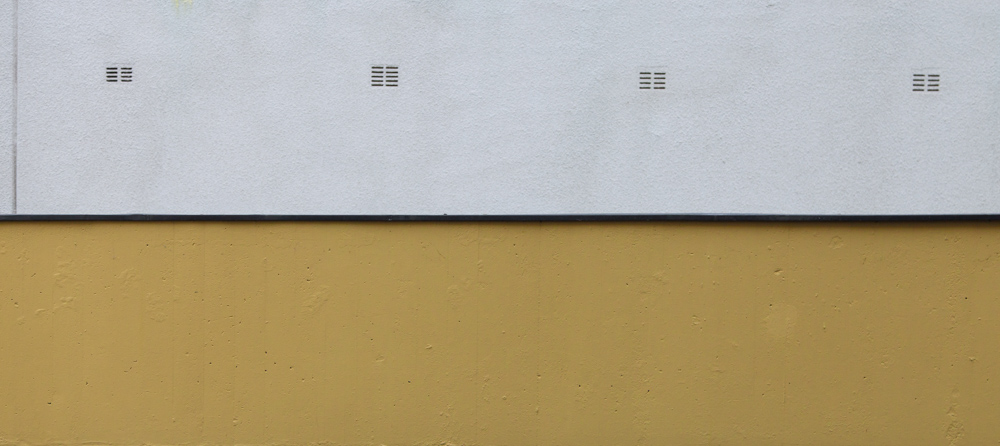 minimalistic horizontal image, seeming two-dimensional, showing a white wall with 4 geometric slits behind a yellow wall