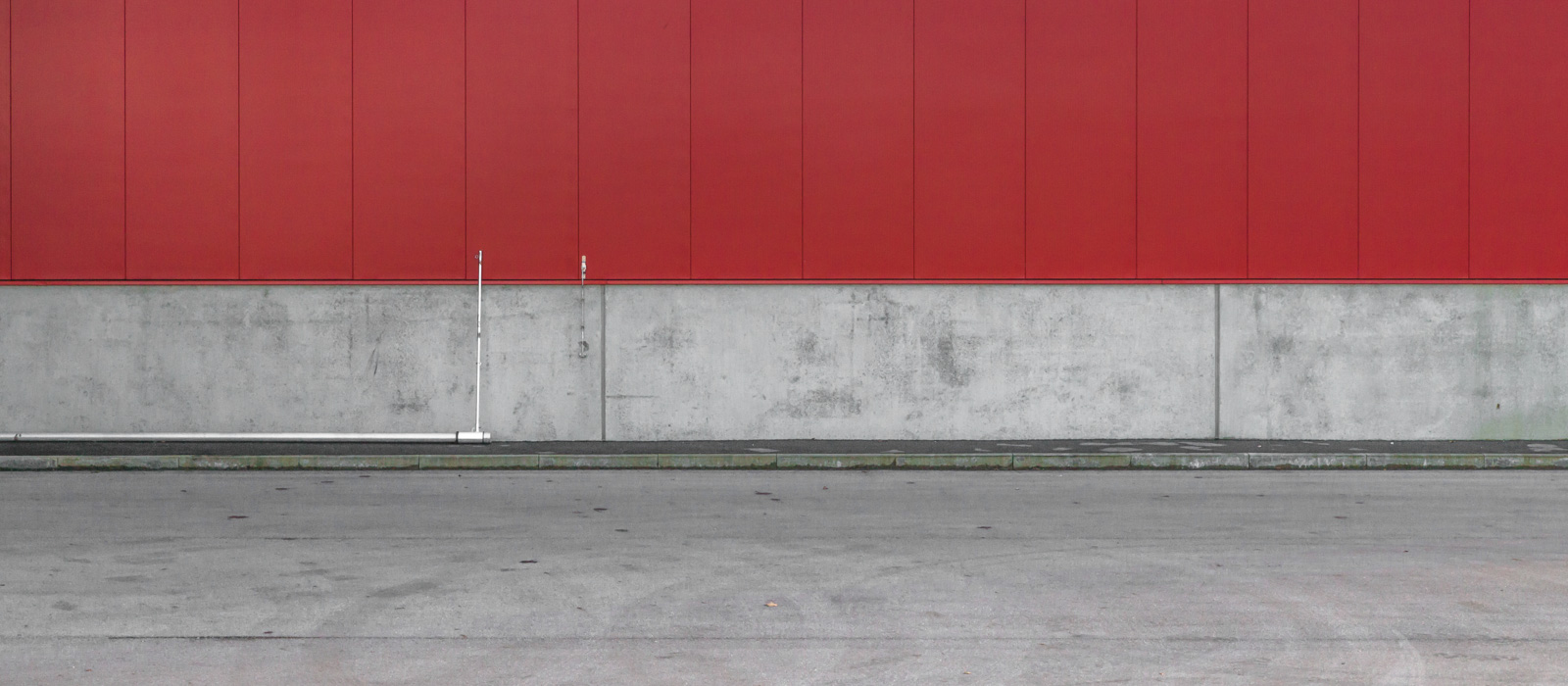 minimalistic horizontal image, seeming two-dimensional, showing a red steel wall with a concrete socket, a pedestrians walk and a street