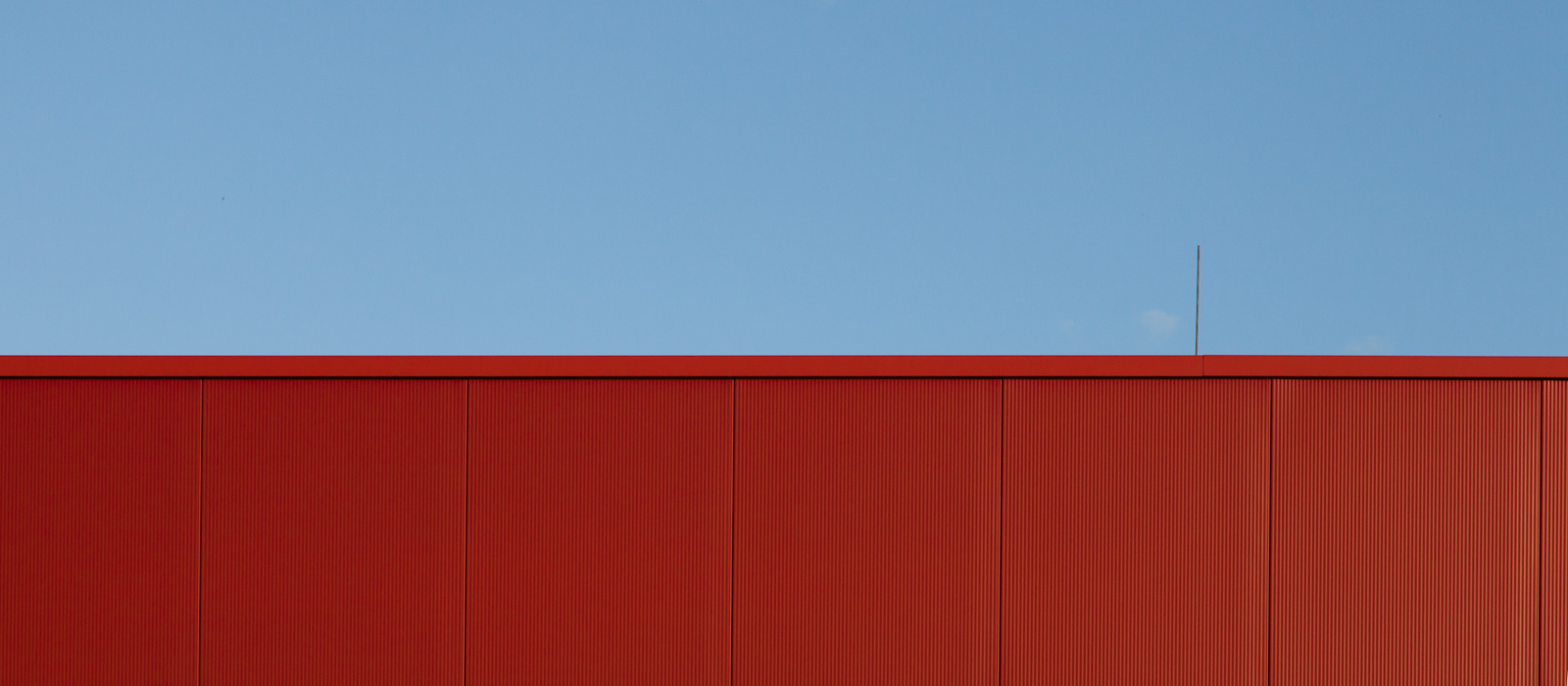 minimalistic horizontal image, seeming two-dimensional, showing a red steel wall with an antenna against the blue sky