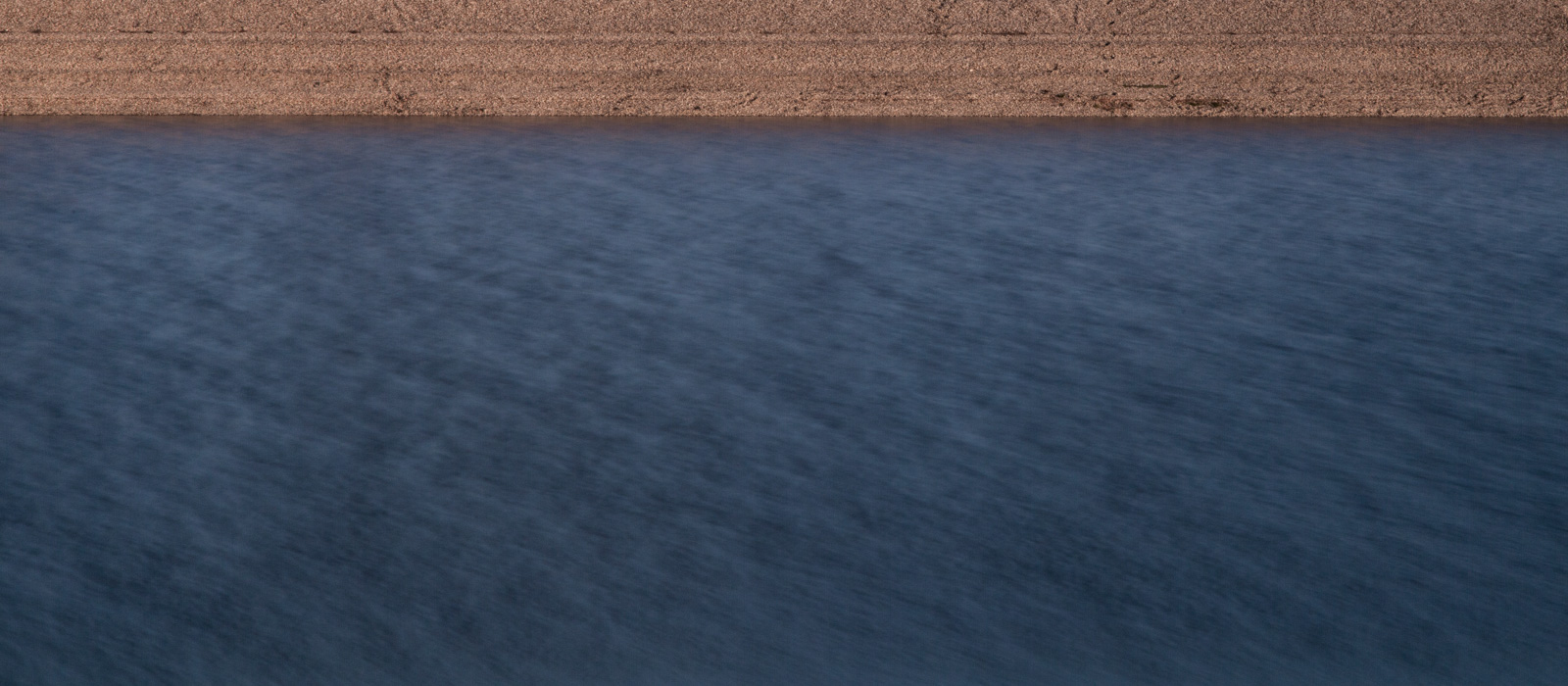 minimalistic horizontal image, seeming two-dimensional, showing a pebble beach and water with a smooth gradient from bright to dark blue and slight blur due to aperture