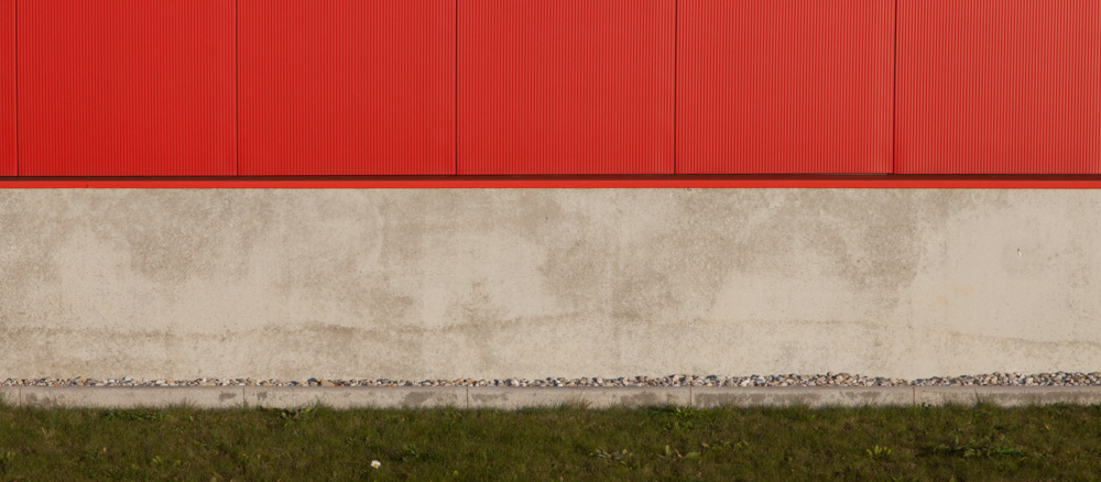 minimalistic horizontal image, seeming two-dimensional, showing a red metal wall with a concrete socket and grass in front of it